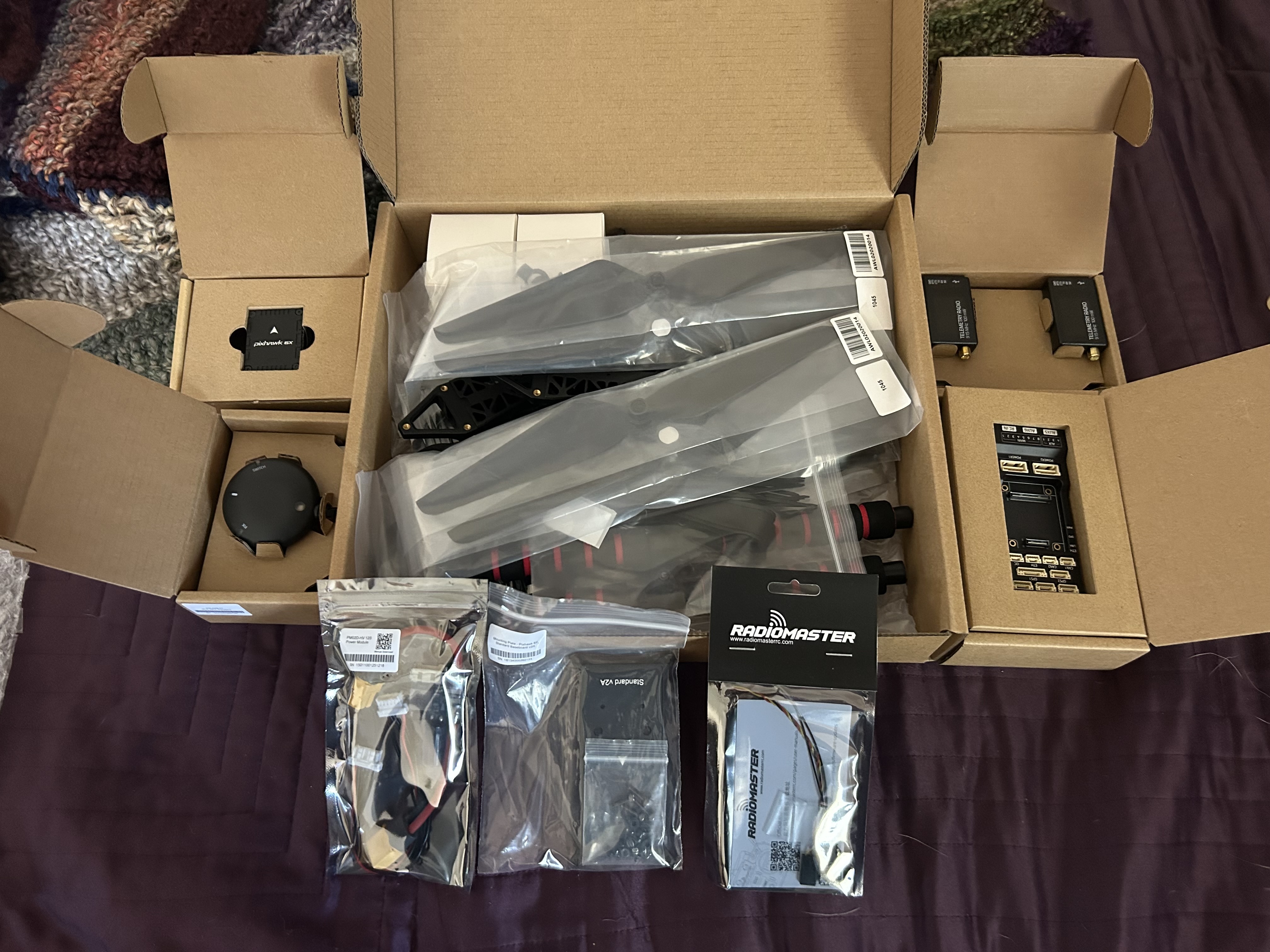 Holybro S500 hardware kit laid out unopened on the bed, including frame parts, props, Pixhawk packaging, telemetry radios, and accessories.